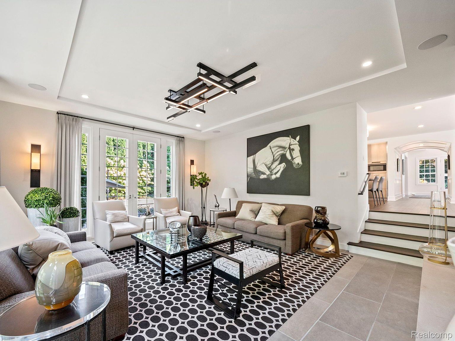 This is an interior shot of a living room featuring a modern design with a black and white patterned rug, neutral-toned sofas, and a unique geometric chandelier. The room is well-lit with natural light streaming through large windows and features a large painting of a horse. The space transitions to a hallway with stairs leading to another level, offering a glimpse into the home's layout.