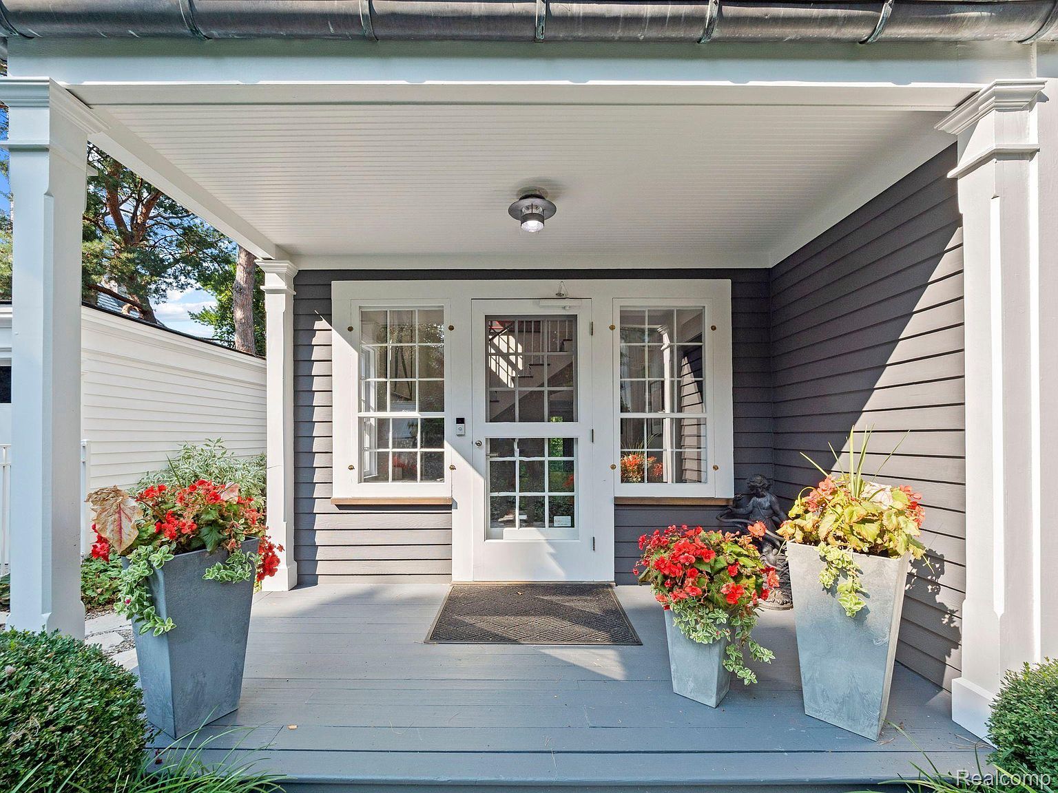 This image showcases a charming entryway with a gray porch, white trim, and a double door with multi-pane windows. Potted plants with vibrant red flowers add a pop of color, enhancing the curb appeal. The porch is well-lit and inviting, creating a warm welcome.