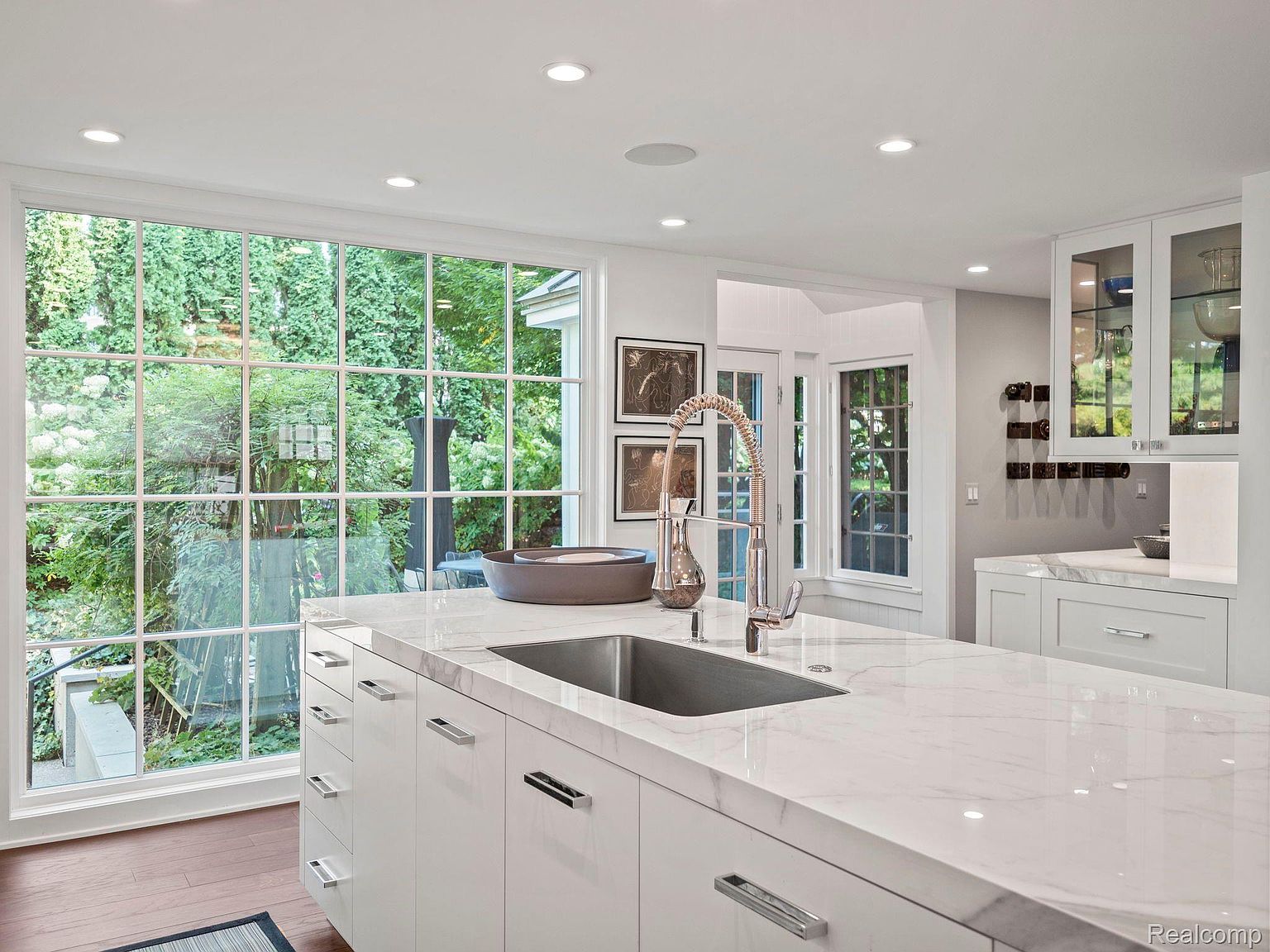 This is a bright and modern kitchen featuring white cabinetry and a large island with a marble countertop. A stainless steel sink and faucet are prominent, and a large window offers a view of the lush greenery outside. The kitchen has a clean and contemporary style, perfect for a modern home.