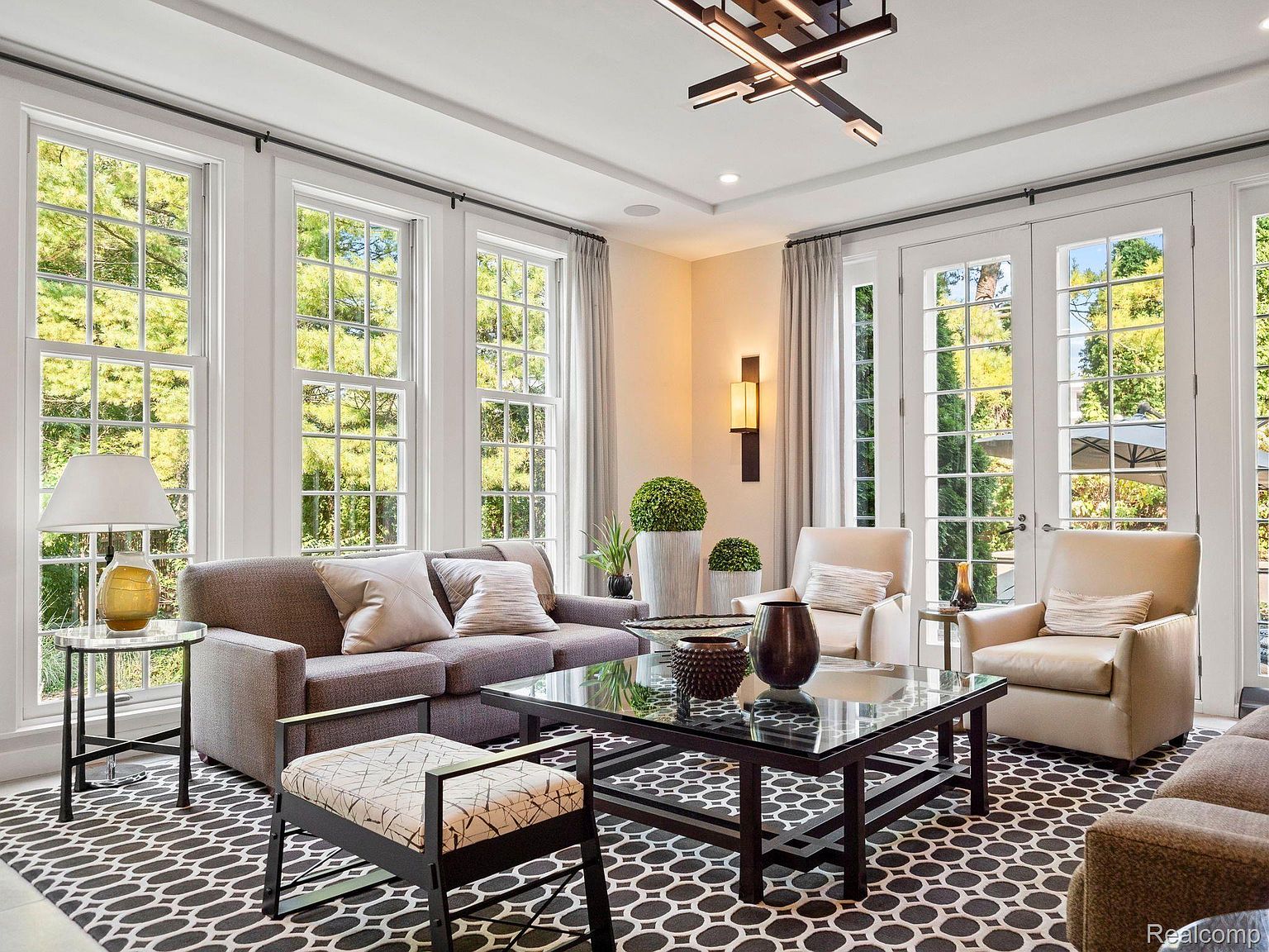 This is a well-lit living room featuring a gray sofa with decorative pillows, two beige armchairs, and a glass-topped coffee table. Large windows provide ample natural light and views of the outdoors. The room is decorated with a black and white patterned rug and modern light fixtures, creating a sophisticated and inviting atmosphere.