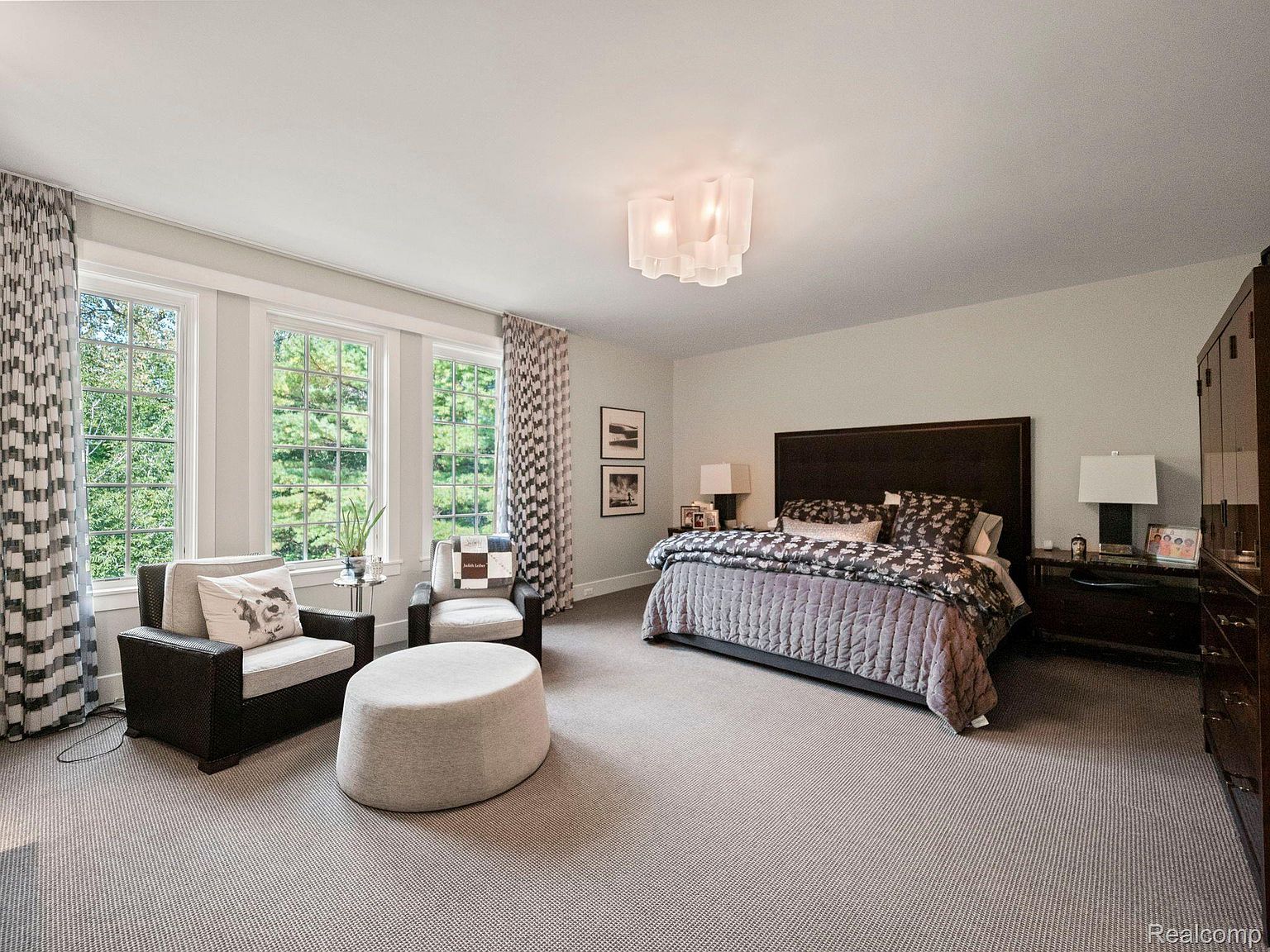This is a spacious primary bedroom featuring a large bed with a dark wood headboard and a patterned comforter. The room includes a seating area with two armchairs and an ottoman, positioned near large windows that provide ample natural light. The color palette is neutral, with light walls and carpeting, creating a serene and inviting atmosphere.