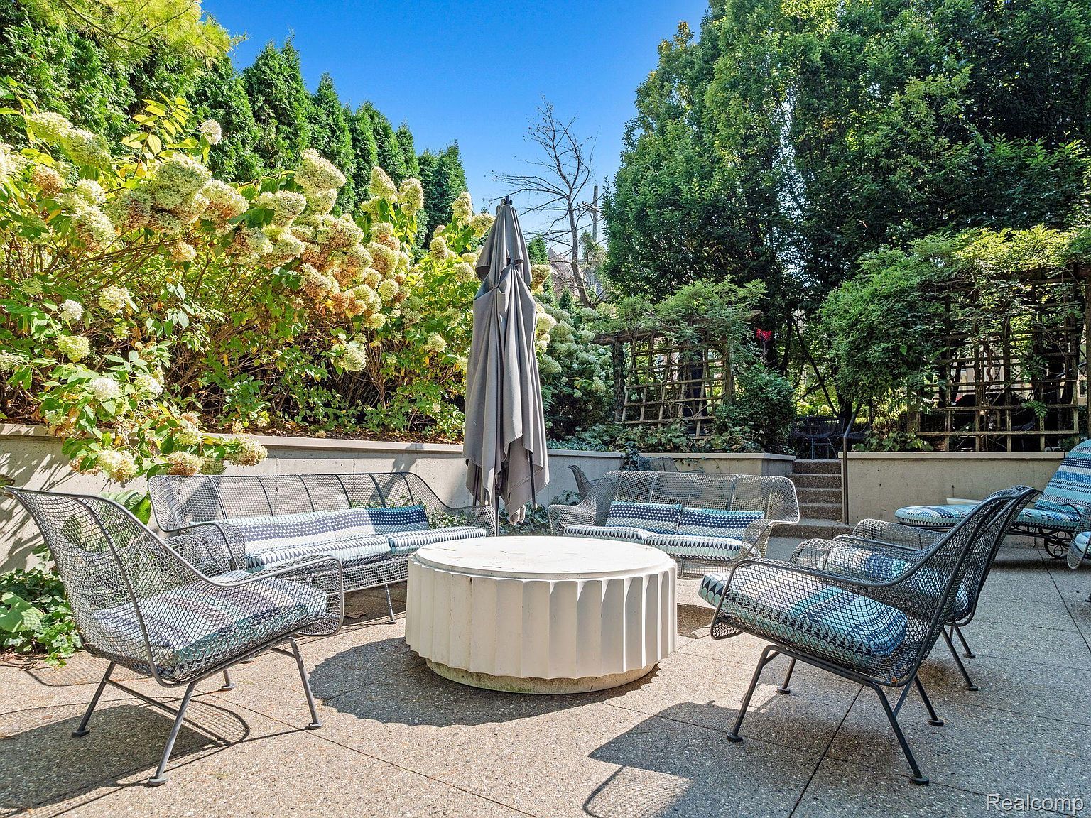 This image showcases a beautifully designed outdoor patio area, perfect for relaxation and entertaining. The space features comfortable seating arrangements with stylish cushions, centered around a unique round table and shaded by a large umbrella. Lush greenery surrounds the patio, creating a private and serene atmosphere.