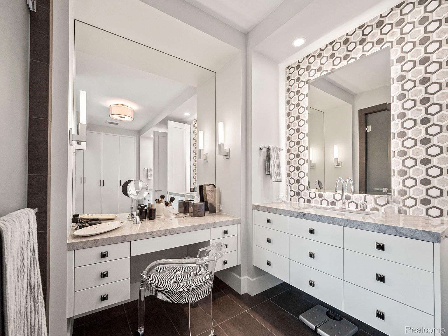 This is a well-lit primary bathroom featuring a vanity area with a large mirror and a makeup station. The bathroom has white cabinetry with dark hardware, a marble countertop, and a decorative tile backsplash around the sink area. The flooring is dark wood, and the overall design is modern and luxurious.