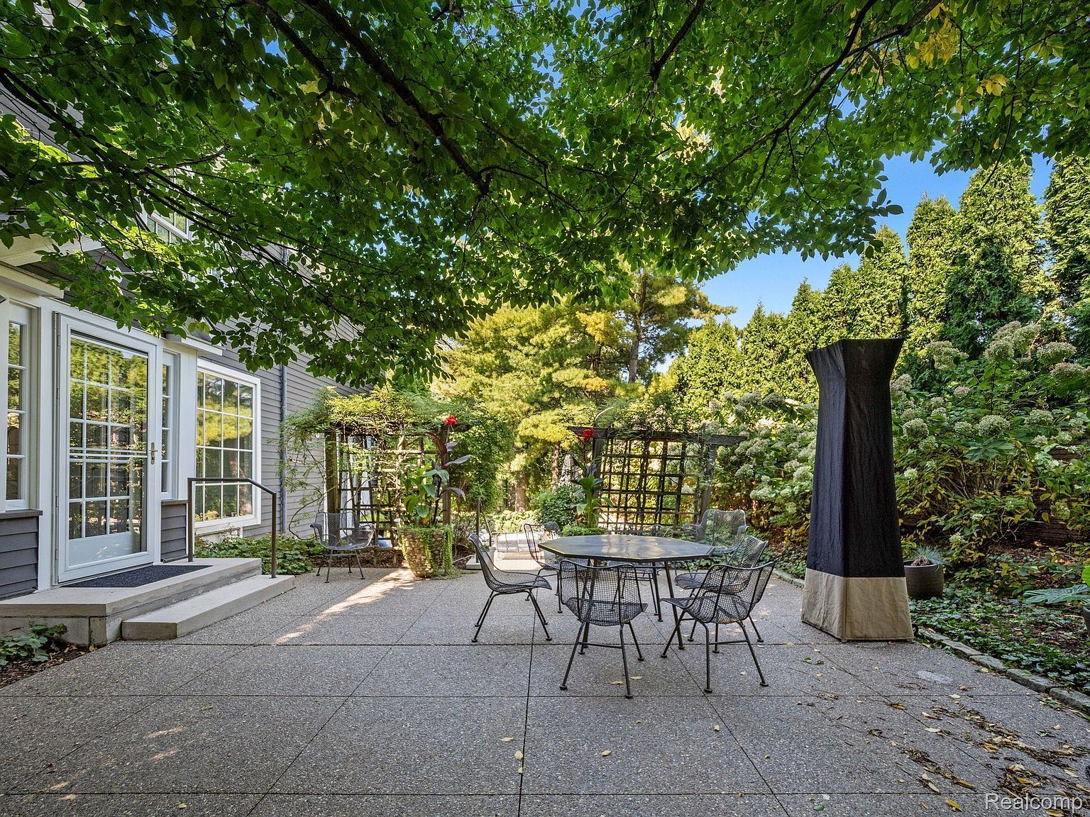 This image showcases a charming outdoor patio area, perfect for relaxation and entertaining. A round metal table with matching chairs sits on a concrete patio, shaded by lush trees. The patio is surrounded by well-maintained landscaping, including trellises with climbing plants and various shrubs, creating a private and inviting outdoor space.