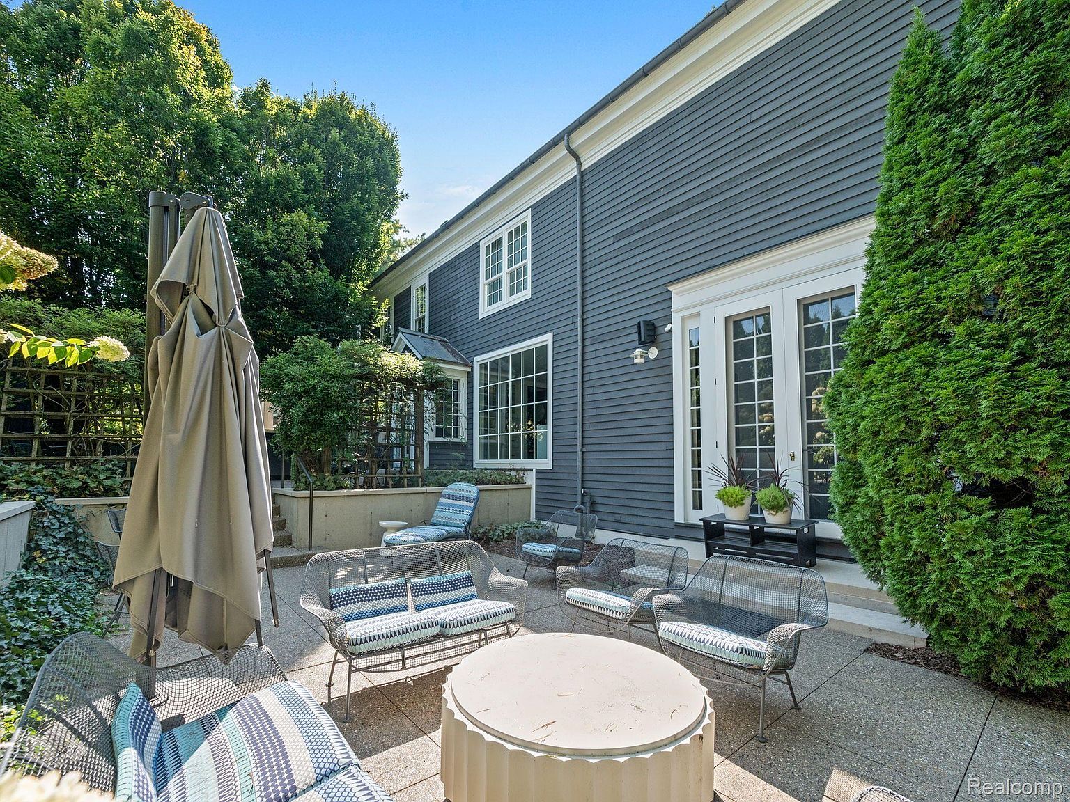 This image showcases a beautifully designed patio area, perfect for outdoor living and entertaining. The space features comfortable seating arrangements with stylish cushions, a central fire pit, and a large umbrella for shade. The patio is adjacent to a well-maintained house with elegant French doors and lush greenery, creating a private and inviting atmosphere.