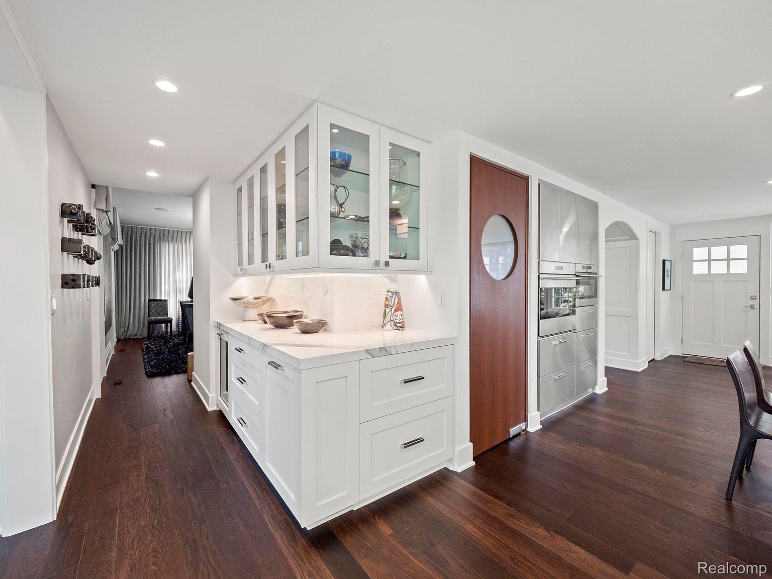 This interior shot showcases a modern kitchen area with dark wood flooring and white cabinetry. The kitchen features a built-in bar area with glass-front cabinets and marble countertops, complemented by stainless steel appliances and a unique circular-windowed door. The space is well-lit with recessed lighting, creating a bright and inviting atmosphere.