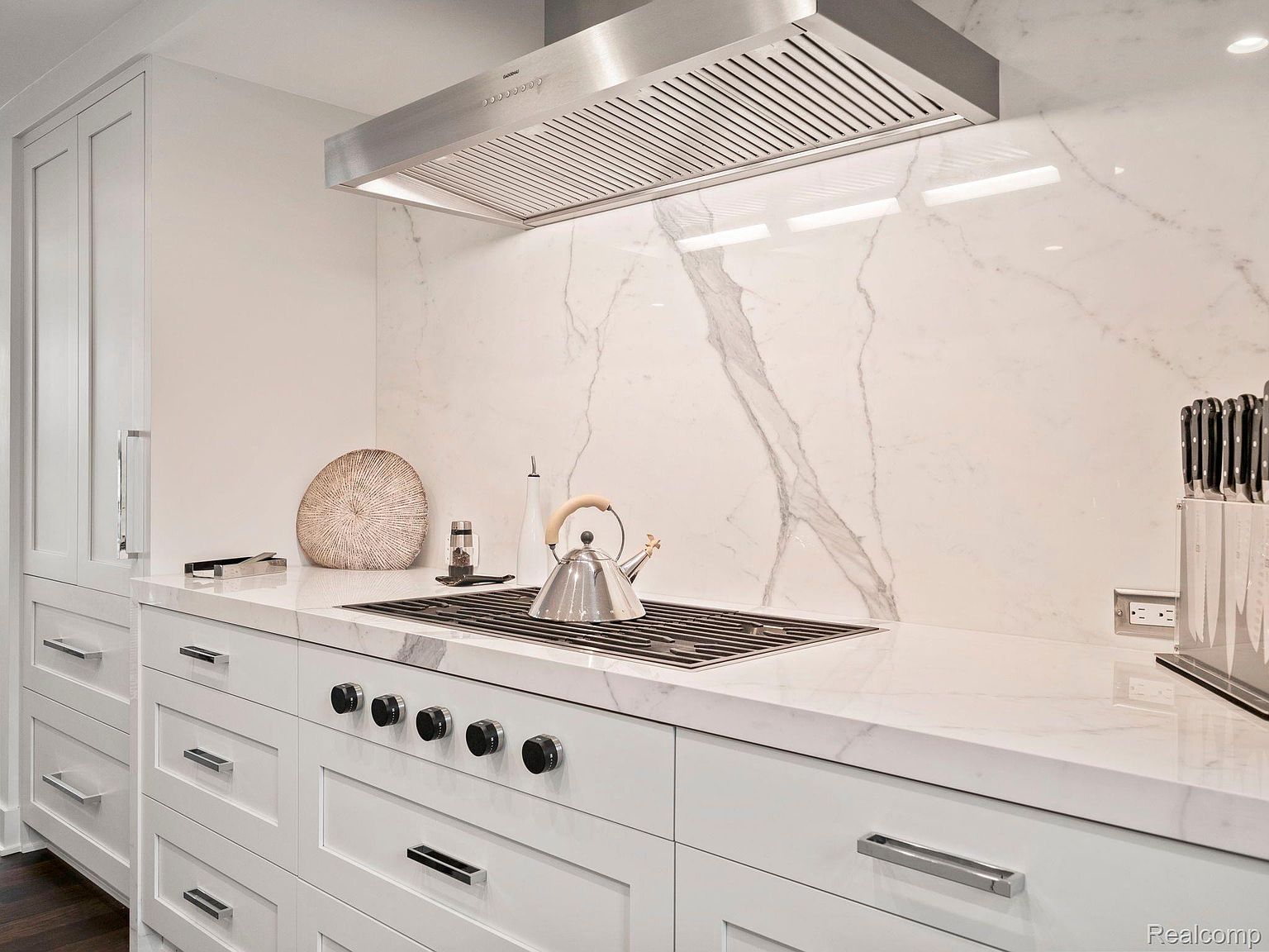 This is a detailed shot of a modern kitchen featuring white cabinetry, marble countertops and backsplash, and stainless steel appliances. The focus is on the stove area, showcasing the sleek design and high-end finishes. The perspective is a medium shot, highlighting the clean lines and elegant style of the kitchen.