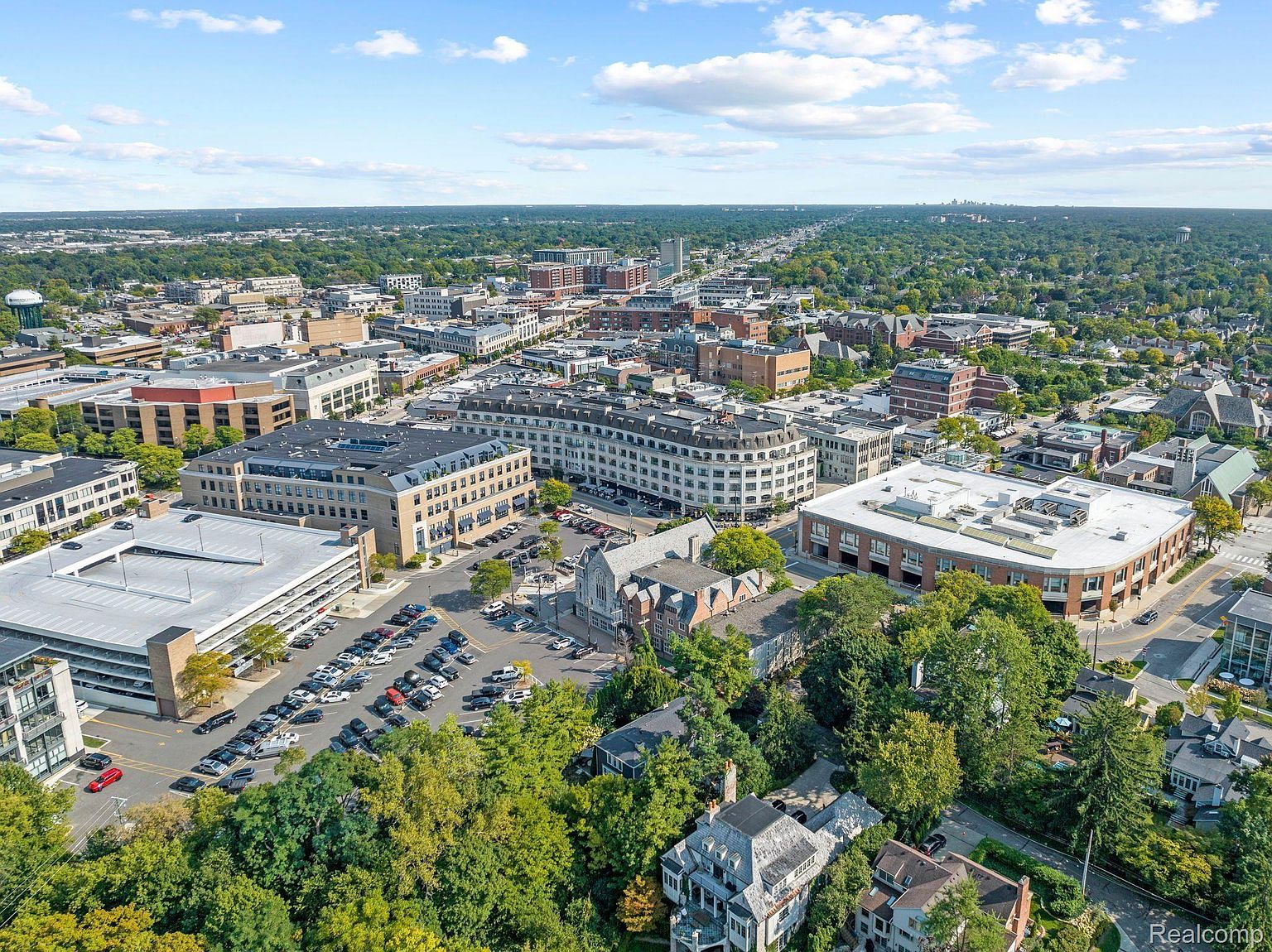 This aerial view showcases a vibrant cityscape with a mix of commercial and residential buildings, surrounded by lush greenery. The architecture varies from modern structures with flat roofs to traditional buildings with pitched roofs, creating a diverse urban landscape. A large parking structure and numerous parked cars indicate a bustling area, while the clear blue sky with scattered clouds adds to the pleasant atmosphere.