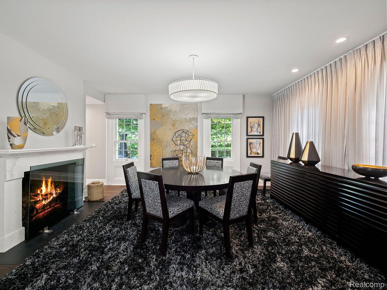 This is an interior shot of a dining room featuring a round dark wood table surrounded by upholstered chairs with dark wood frames. A modern chandelier hangs above the table, and a dark wood sideboard with decorative vases sits against the wall. The room is decorated in neutral tones with a fireplace on the left and a large abstract painting visible in the background, creating a sophisticated and inviting atmosphere.