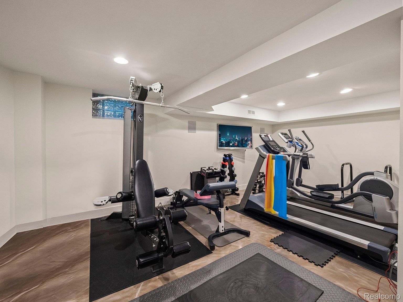 This is an interior shot of a home gym featuring a multi-station weight machine, a treadmill, and an elliptical machine. The room has a modern feel with recessed lighting, neutral-colored walls, and a television mounted on the wall. The floor is covered with a combination of rubber mats and a polished surface, creating a dedicated workout space.