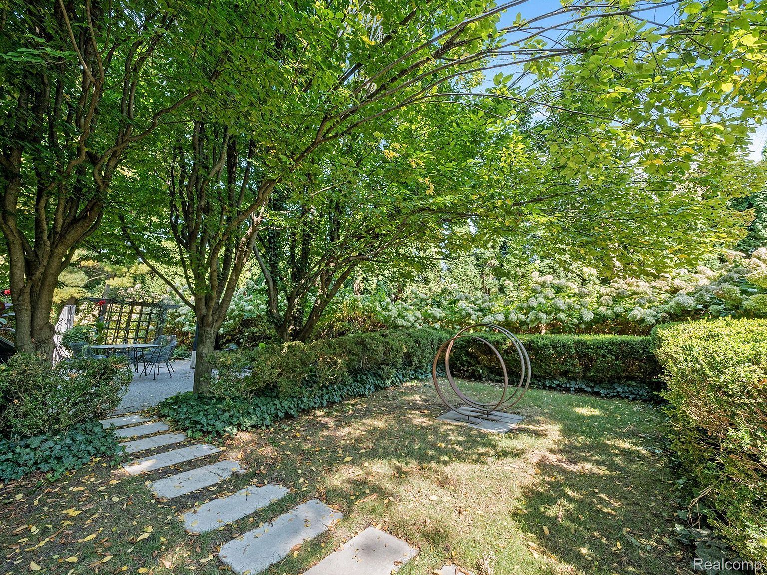 This image showcases a beautifully landscaped yard or garden, featuring lush greenery, mature trees providing ample shade, and a stone pathway. A unique circular metal sculpture adds an artistic touch to the space. The overall impression is one of tranquility and natural beauty, making it an appealing outdoor retreat.