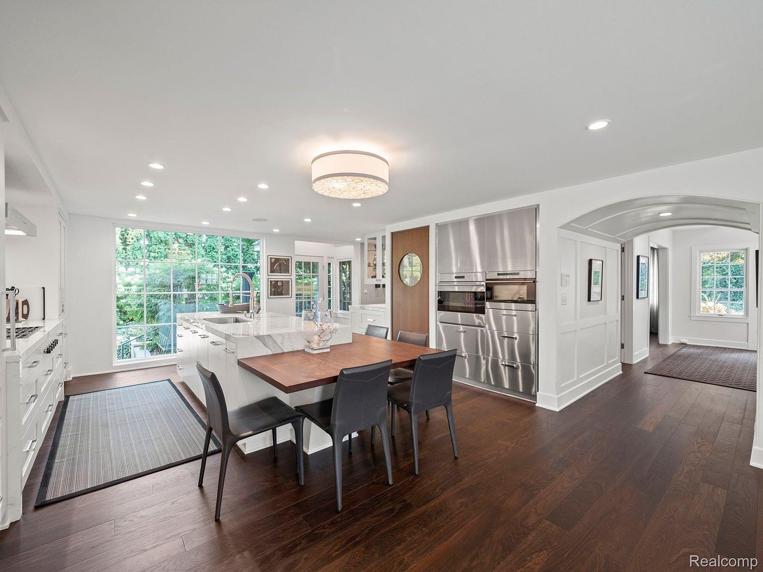 This is a bright and spacious kitchen featuring dark hardwood floors, white cabinetry, and stainless steel appliances. A large window provides ample natural light and a view of the outdoors. A dining table with modern chairs sits adjacent to the kitchen island, creating an inviting space for meals.