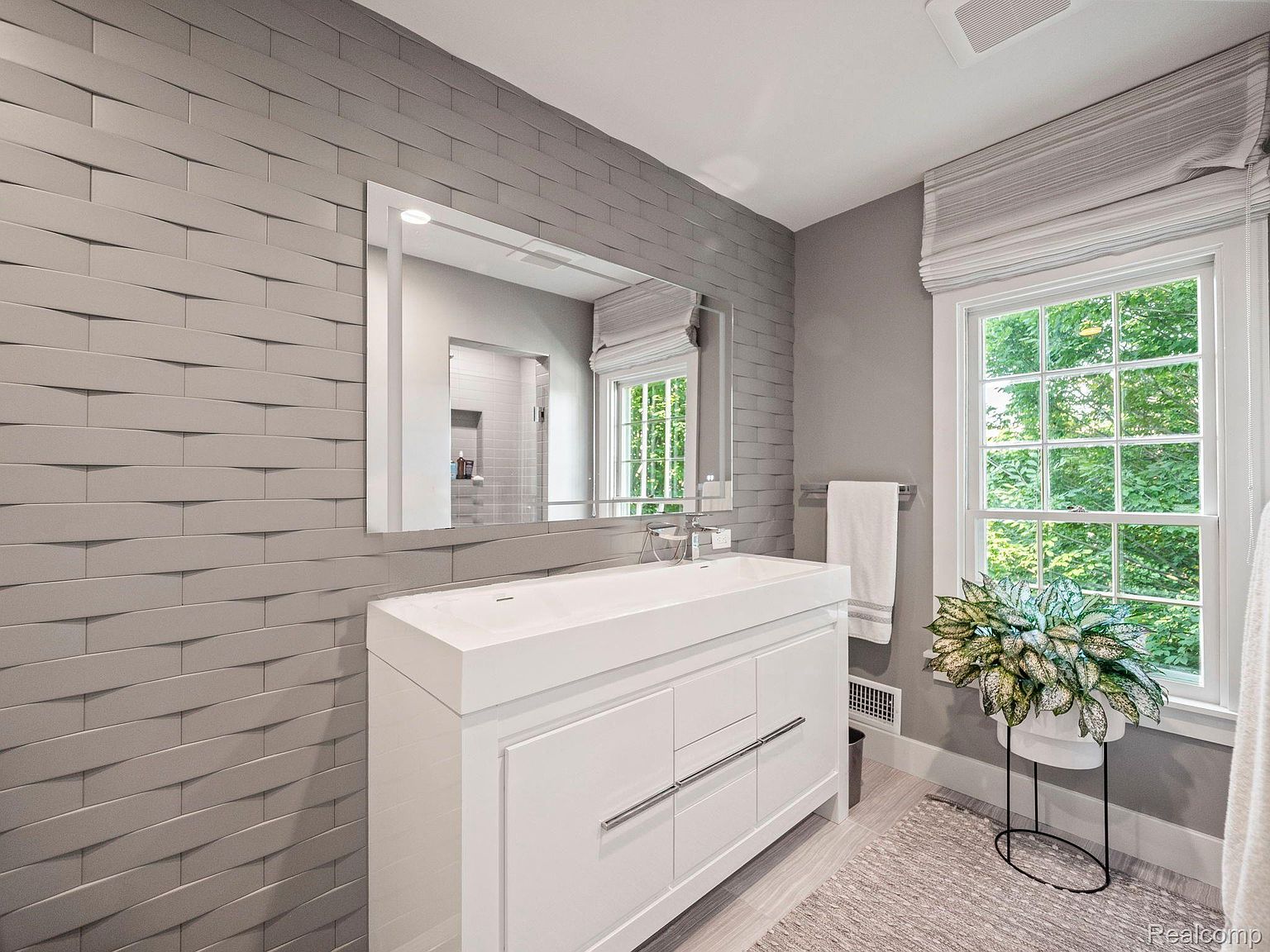 This is a modern bathroom featuring a white vanity with a sleek countertop and integrated sink. A large mirror hangs above the vanity, reflecting the window and natural light. The wall is adorned with textured gray tiles, adding depth and visual interest to the space.
