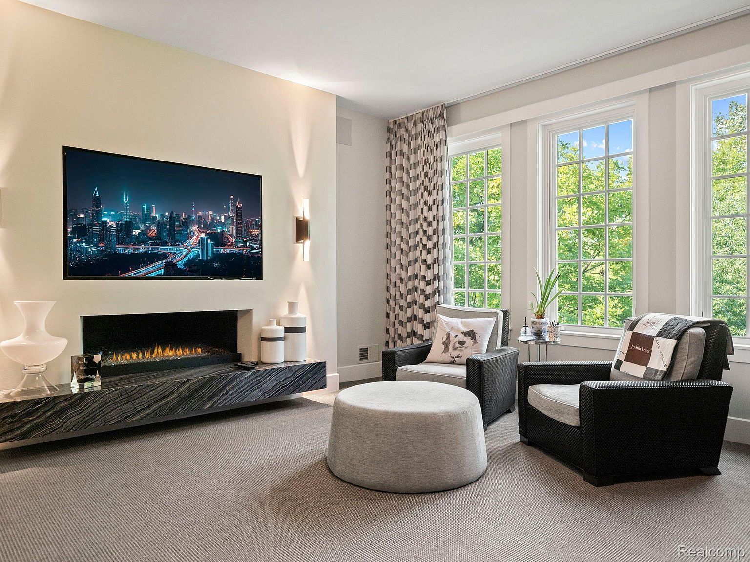 This is an interior shot of a living room featuring a modern fireplace with a large television mounted above it. Two armchairs flank a round ottoman, positioned near large windows that offer a view of lush greenery. The room is decorated in neutral tones, creating a serene and inviting atmosphere.