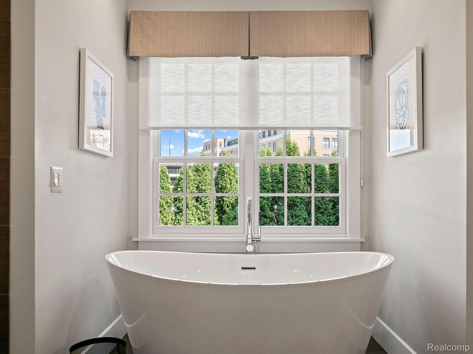 This is a bright and airy primary bathroom featuring a freestanding white bathtub positioned in front of a large window. The window provides a view of lush greenery and nearby buildings, while the room is decorated with framed artwork on the walls. The overall aesthetic is clean, modern, and spa-like, creating a relaxing atmosphere.
