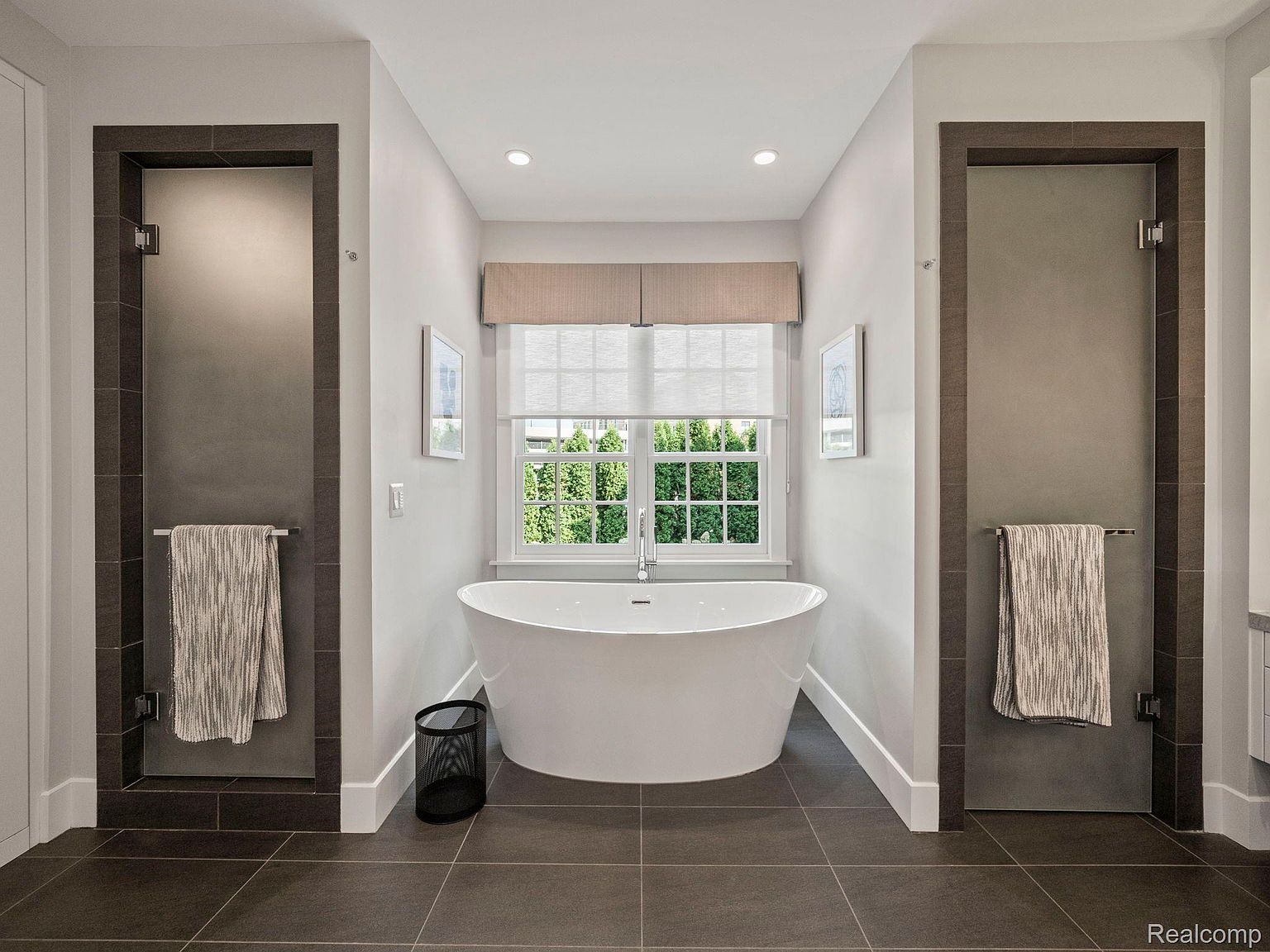 This is a well-lit primary bathroom featuring a freestanding white bathtub positioned in front of a window with natural light streaming in. The bathroom has a modern design with gray tile flooring, neutral wall colors, and dark-framed shower entrances on either side of the tub. The overall impression is clean, serene, and luxurious.