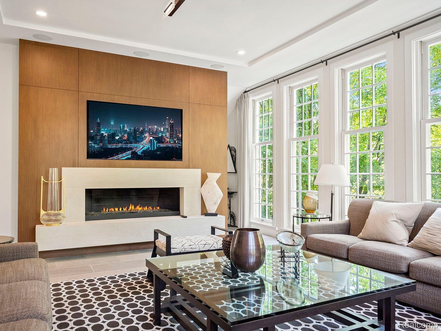This is a well-lit living room featuring a modern fireplace with a large television mounted above it. The room has large windows offering natural light and views of the outdoors. The decor includes a glass coffee table, vases, and a patterned rug, creating a stylish and comfortable atmosphere.