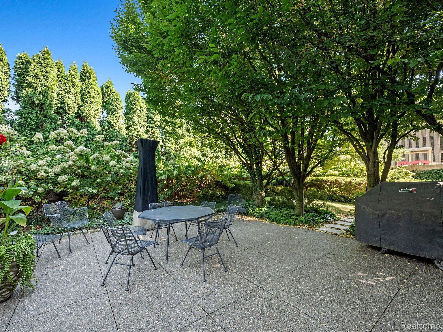 This image showcases a charming outdoor patio area, perfect for relaxation and entertaining. The space features a stone-paved patio with a metal table and chairs set, surrounded by lush greenery, including trees, bushes, and flowering plants. A grill with a cover is also visible, suggesting outdoor cooking possibilities.