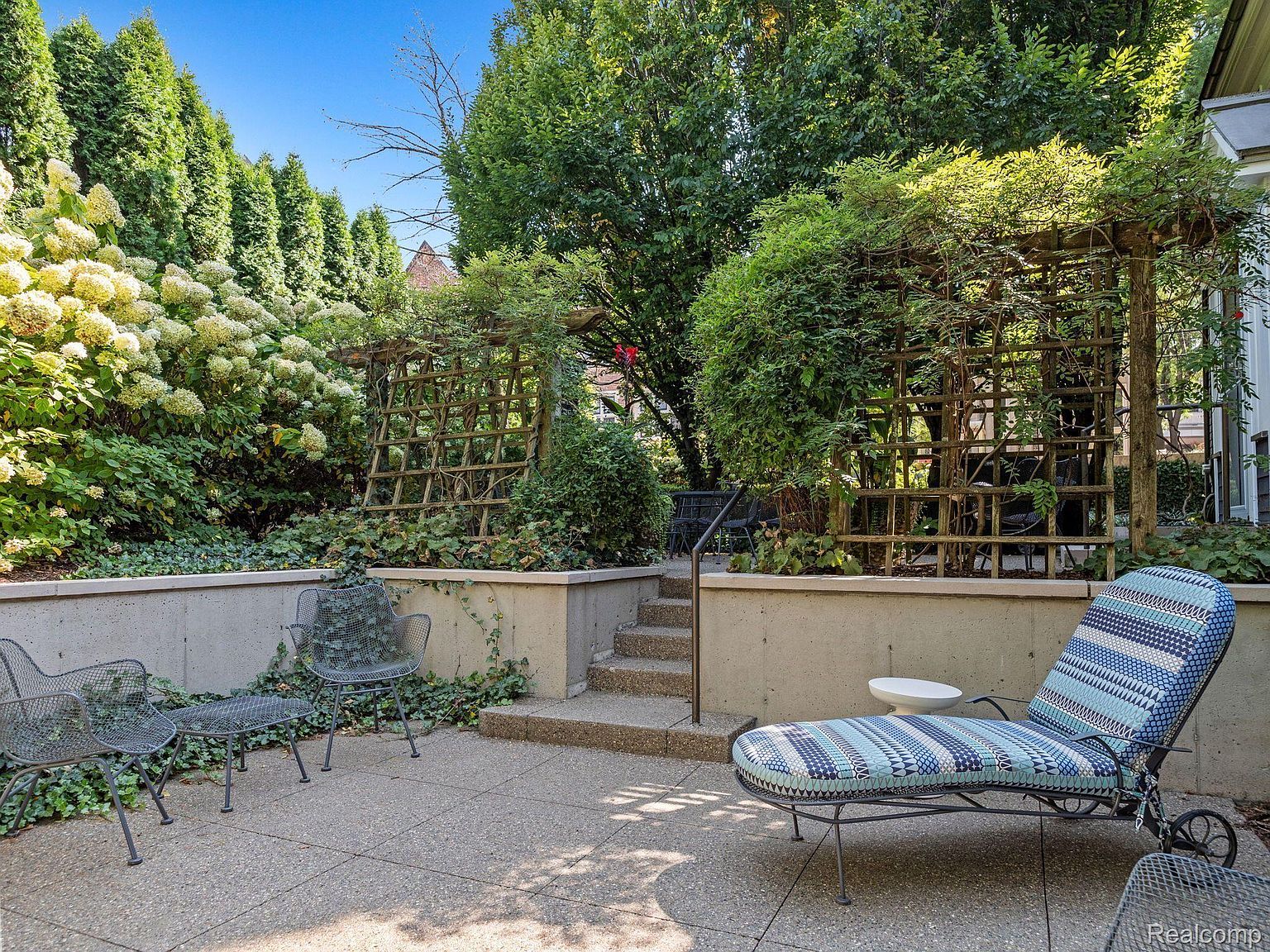 This image showcases a charming outdoor patio area, featuring a mix of seating options including metal chairs and a cushioned chaise lounge. The patio is surrounded by lush greenery, including tall hedges, climbing plants on trellises, and ivy-covered walls, creating a private and serene atmosphere. The space is paved with concrete, and a small set of stairs leads to a higher level of the garden.