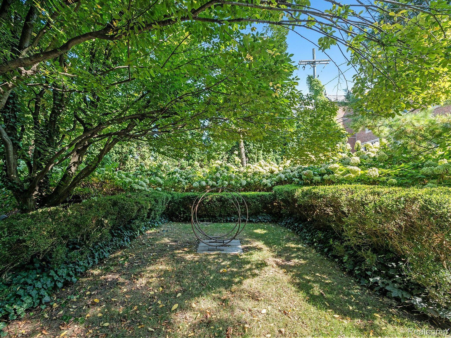 This image showcases a beautifully maintained garden with lush greenery, including manicured hedges and flowering plants. A circular metal sculpture sits on a well-kept lawn, adding an artistic touch. The garden appears serene and inviting, suggesting a peaceful outdoor space for relaxation and enjoyment.