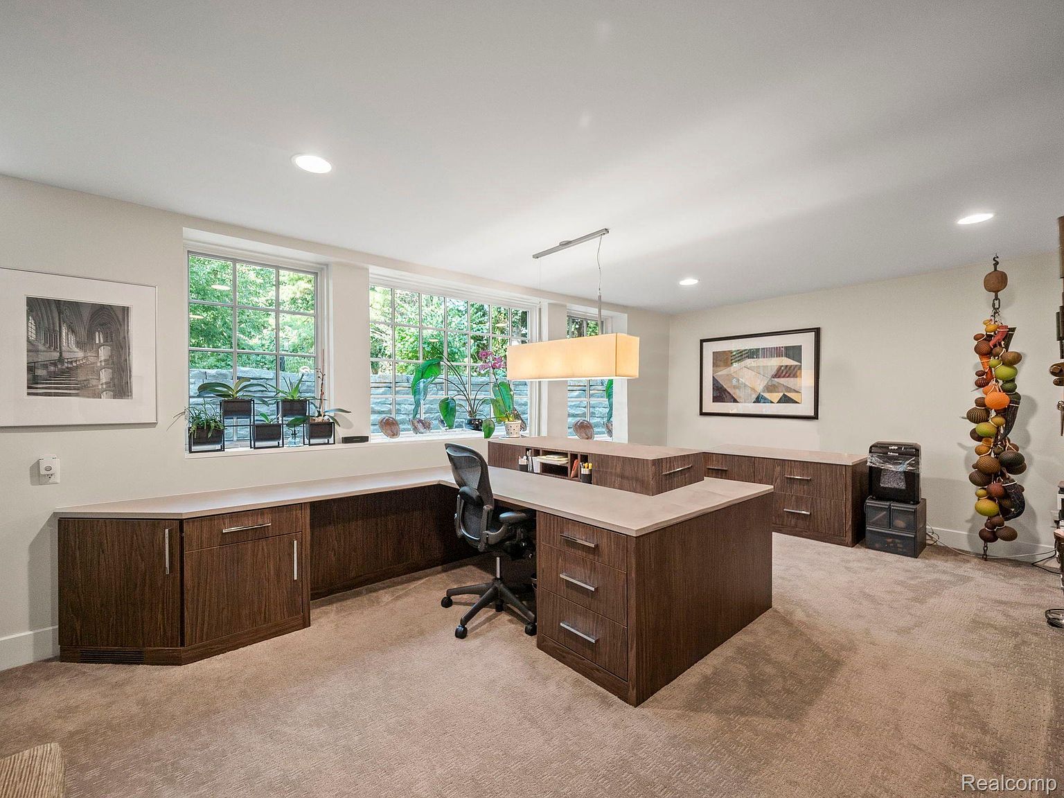 This is an interior shot of a well-organized office space. The room features a large, custom-built desk with ample storage, complemented by a neutral color palette and natural light streaming through the windows. The overall impression is one of functionality and sophistication, ideal for a productive work environment.