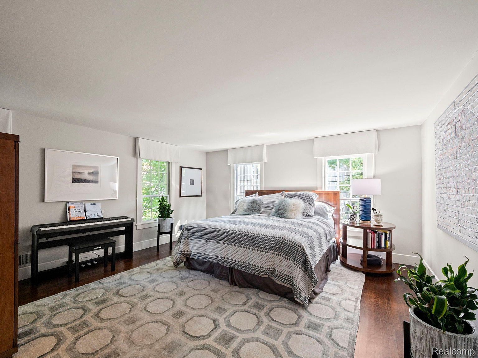 This is a spacious primary bedroom featuring a large bed with a patterned comforter and decorative pillows. The room is well-lit with natural light from multiple windows, which are adorned with simple white valances. A piano sits against one wall, and a round side table with a lamp and books adds a cozy touch. The room is decorated in neutral tones with a large area rug, creating a serene and inviting atmosphere.