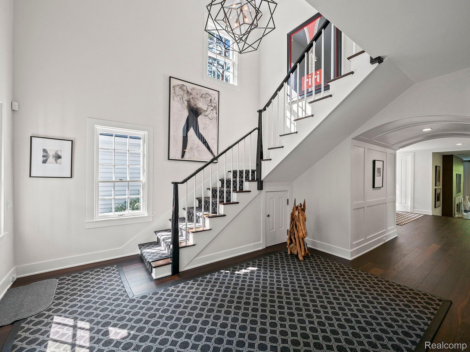 This interior shot showcases a grand hallway and staircase, featuring a dark wood floor, a patterned area rug, and white walls adorned with artwork. The staircase has a black railing with white spindles and a patterned runner. Natural light streams in through windows, highlighting the architectural details and creating a bright and inviting space.