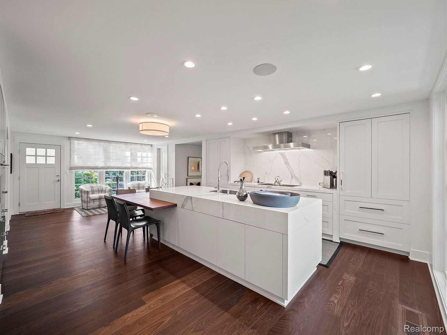 This is a bright and spacious kitchen featuring a large island with a waterfall countertop and seating. The kitchen is equipped with modern stainless steel appliances and white cabinetry, complemented by a marble backsplash. Dark hardwood floors add warmth to the space, and natural light floods in through a nearby window.