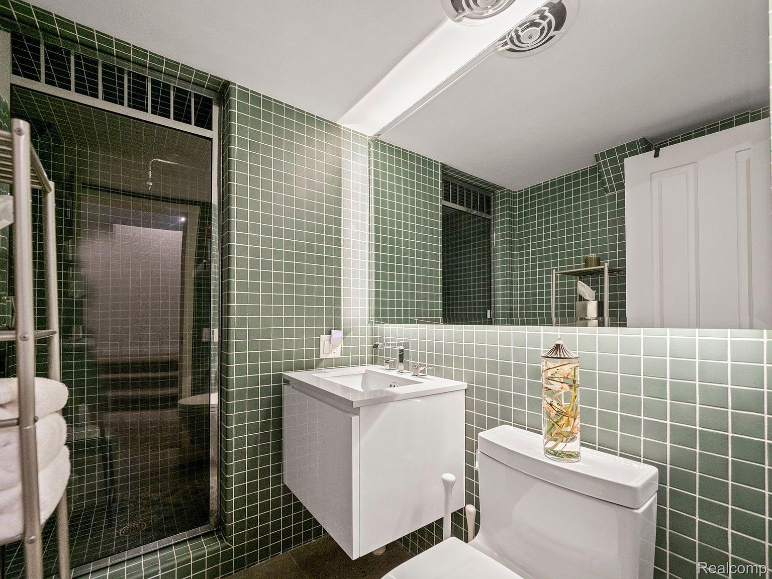 This is a bathroom featuring green square tiles on the walls and floor, creating a retro yet modern aesthetic. The bathroom includes a glass-enclosed shower, a floating white vanity with a sink, and a toilet with a decorative glass vase on top. A large mirror reflects the space, enhancing the sense of openness.
