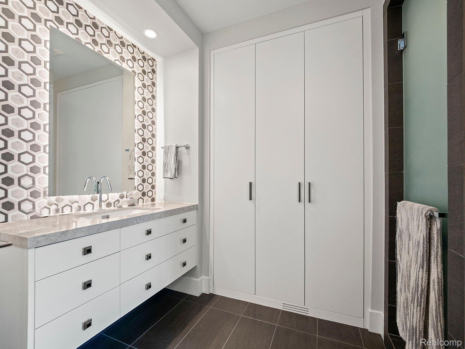 This is a well-lit primary bathroom featuring a modern vanity with white drawers and a light-colored countertop. A decorative mirror with a geometric tile border hangs above the vanity. To the right, there is a tall, white storage cabinet with sleek, dark handles, and a glimpse of a shower area with a towel hanging on a bar.
