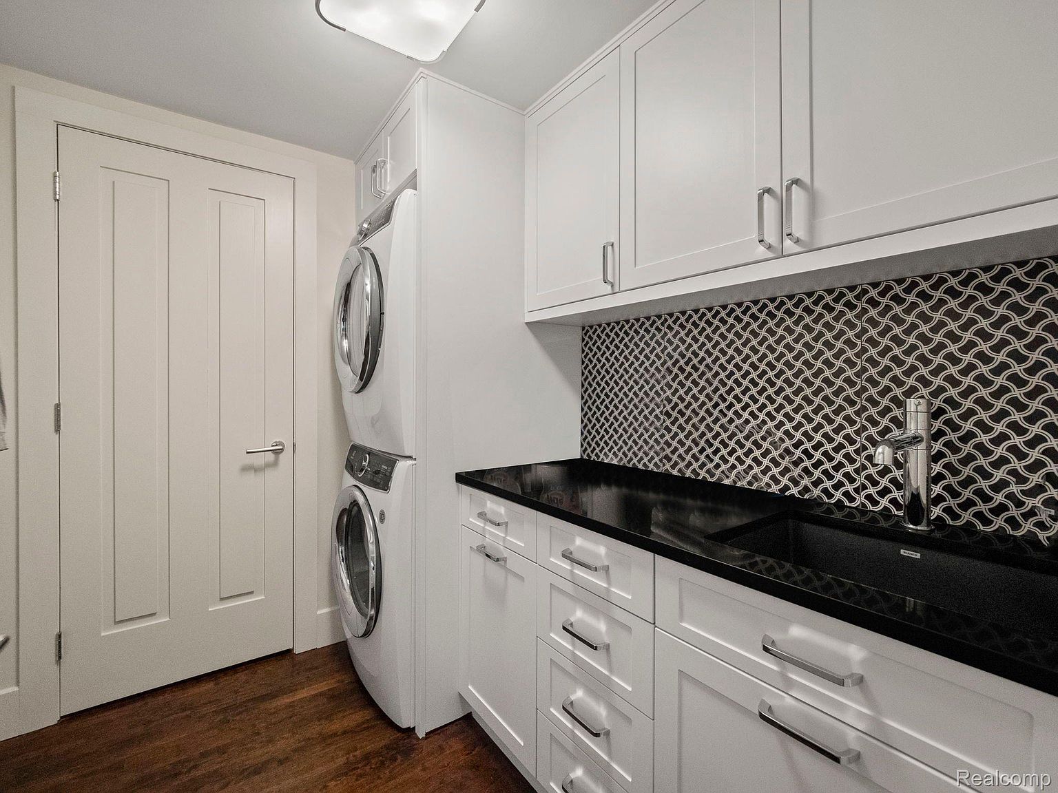 This is a well-organized laundry room featuring a stacked washer and dryer unit, white cabinetry with sleek hardware, and a black countertop with a sink. The backsplash is a decorative mosaic tile, adding a touch of elegance to the space. The room is clean and functional, designed for efficient laundry tasks.