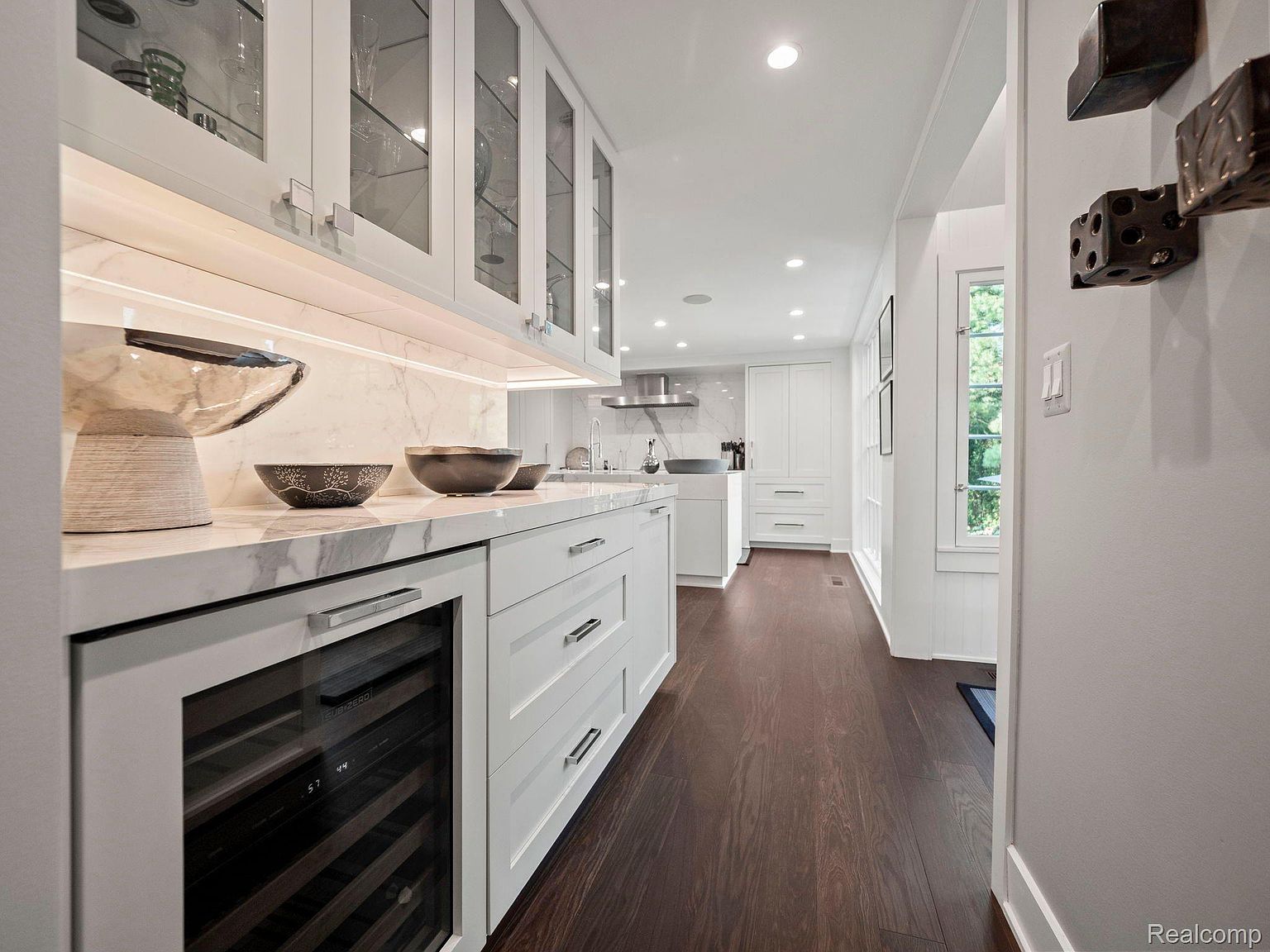 This is a bright, modern kitchen featuring white cabinetry with glass fronts and marble countertops. A built-in wine fridge is visible, and the dark hardwood floors add a touch of warmth. The kitchen appears spacious and well-lit, creating an inviting atmosphere.