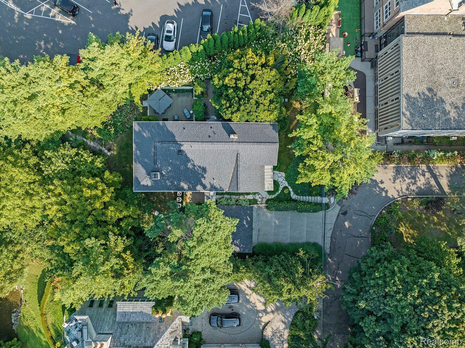 This aerial shot showcases a well-maintained property featuring a house with a dark gray roof, surrounded by lush greenery and mature trees. A paved driveway leads to the house, and a patio area with outdoor furniture is visible. The image highlights the property's landscaping, privacy, and overall curb appeal.