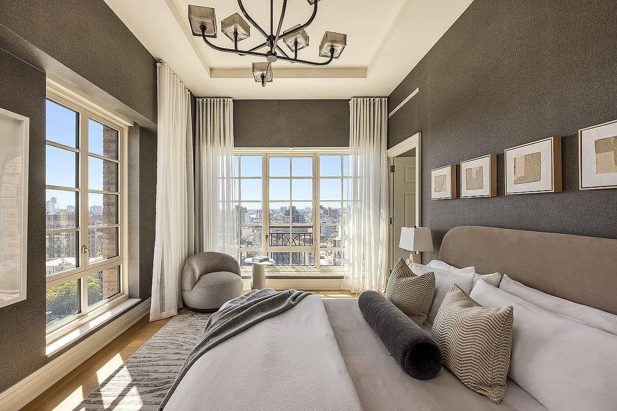 This elegant primary bedroom features dark, textured wallpaper and a neutral color palette, creating a sophisticated and serene atmosphere. A large bed with plush pillows and a bolster cushion serves as the focal point, complemented by a modern chandelier and a cozy armchair positioned near the expansive windows that offer a city view. The room is bathed in natural light, highlighting the clean lines and refined decor, including a series of framed art pieces above the headboard.