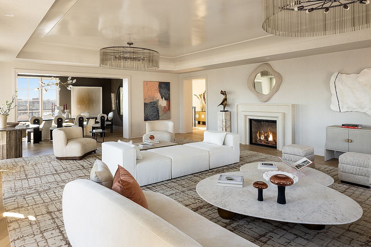 This sophisticated living room features a neutral color palette with plush, curved seating arranged around a large, organic-shaped stone coffee table. A fireplace with a classic mantel serves as a focal point, complemented by a decorative mirror and abstract wall art. The space is illuminated by two large, circular tiered chandeliers, creating an elegant and airy atmosphere that flows seamlessly into the adjacent dining area.