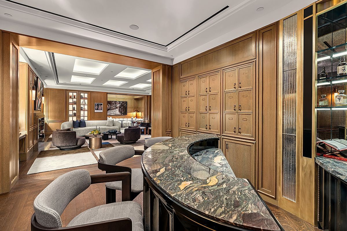 This sophisticated interior space features a foreground bar area with a curved, patterned stone countertop and two upholstered chairs. Beyond the bar, the room opens into a spacious living area characterized by warm wood-paneled walls, a neutral color palette, and plush seating arrangements. The perspective captures a seamless transition between the intimate bar setting and the expansive, well-lit living room, highlighting a refined and luxurious aesthetic.