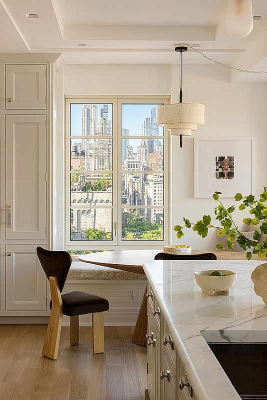This bright and sophisticated kitchen features a large island with a stone countertop and a built-in sink, overlooking a cozy dining nook. A window with a view of the city skyline serves as a focal point, complemented by a tiered pendant light and a unique wooden chair. The space is finished with light wood flooring and neutral cabinetry, creating a clean and elegant atmosphere.