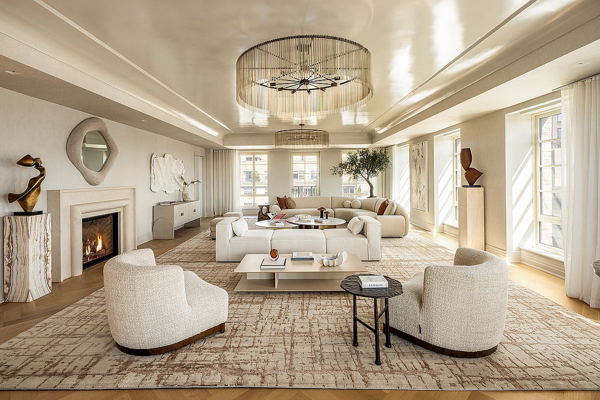 This expansive, light-filled living room features a sophisticated neutral palette with plush, curved seating arrangements centered around a low-profile coffee table. A prominent stone fireplace with a sculptural art piece above serves as a focal point, while large windows allow natural light to flood the space. The room is anchored by a textured area rug and illuminated by a striking tiered chandelier, creating an atmosphere of refined, contemporary luxury.