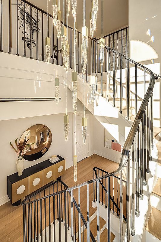 This elegant entryway features a dramatic, multi-tiered crystal chandelier that cascades down the center of a grand staircase. The space is defined by a sleek, dark metal railing, light wood flooring, and a modern console table paired with a circular mirror. The perspective from the upper landing highlights the architectural flow and the bright, airy atmosphere created by the natural light.