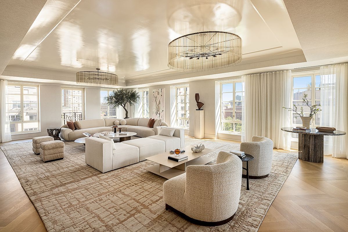 This expansive, light-filled living room features a sophisticated neutral palette with a large, curved sectional sofa and plush armchairs arranged on a textured area rug. The space is anchored by two striking, circular tiered chandeliers and tall windows that provide abundant natural light and city views. The room is finished with light wood flooring in a herringbone pattern and minimalist decor, creating an atmosphere of refined, modern luxury.