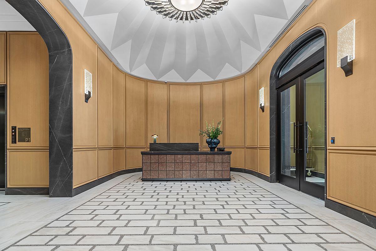 This elegant lobby or reception area features a curved wall paneled in light wood, accented by dark stone trim and arched doorways. A central reception desk with a textured, dark-toned front sits on a patterned stone floor, while a dramatic, faceted ceiling design draws the eye upward to a modern chandelier. The space conveys a sophisticated, high-end atmosphere suitable for a luxury residential building or boutique hotel.