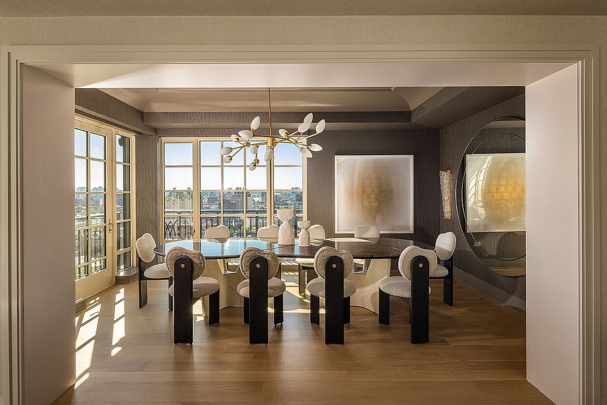 This sophisticated dining room features a large, dark oval table surrounded by modern chairs with unique, sculptural black frames and textured light-colored upholstery. A striking, multi-armed chandelier hangs above the table, while large windows provide an expansive view of the city skyline. The room is framed by a wide, clean-lined doorway and features dark, textured wall coverings that contrast with the light wood flooring.