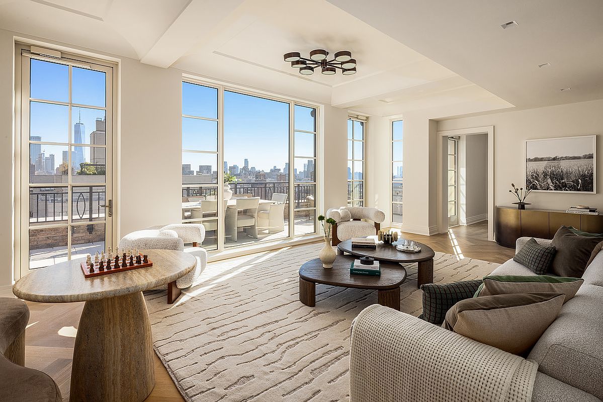 This sophisticated living room features a neutral color palette, plush textured seating, and a large area rug that anchors the space. Large windows provide abundant natural light and offer a clear view of the city skyline and an adjacent outdoor terrace. The room is furnished with organic-shaped coffee tables and a chess set, creating an inviting and elegant atmosphere.
