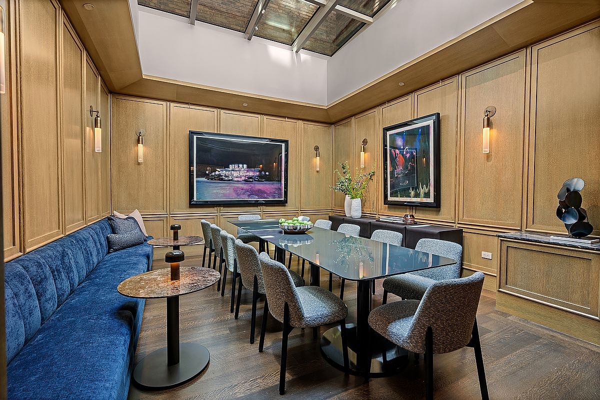 This sophisticated dining room features wood-paneled walls and a long, dark glass-topped table surrounded by modern upholstered chairs. A plush blue velvet banquette runs along one wall, complemented by two small circular side tables, while a skylight above provides natural illumination. The space is accented by framed artwork and wall-mounted light fixtures, creating an elegant and intimate atmosphere.