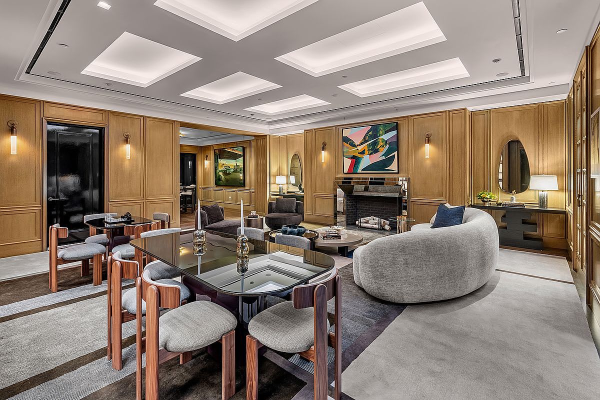 This sophisticated living and dining area features warm wood-paneled walls and a recessed tray ceiling with integrated lighting. The space is furnished with a curved, textured sofa, a glass-topped dining table, and modern wood-framed chairs, all arranged on a layered area rug. A fireplace with a reflective surround serves as a focal point, complemented by abstract artwork and elegant wall sconces.