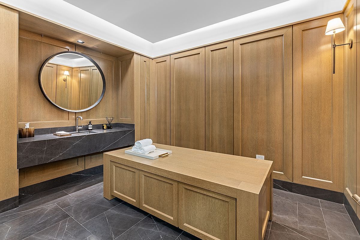 This sophisticated spa-like bathroom features extensive light wood paneling that creates a warm, cohesive aesthetic. A dark stone vanity with a circular mirror and a central massage or treatment table with folded towels define the space, while dark stone flooring adds a grounded contrast. The room is illuminated by recessed lighting and a wall-mounted sconce, offering a serene and luxurious atmosphere.