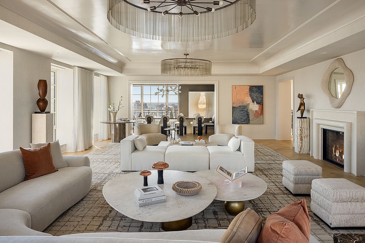 This expansive, sophisticated living room features a neutral color palette with layered textures, including a large patterned area rug and plush white seating. The space is anchored by a central stone fireplace with a sculptural mirror above and is illuminated by two large, tiered circular chandeliers. Through the open doorway, a glimpse of a dining area and a view of the city skyline beyond large windows adds depth to this elegant, open-concept layout.