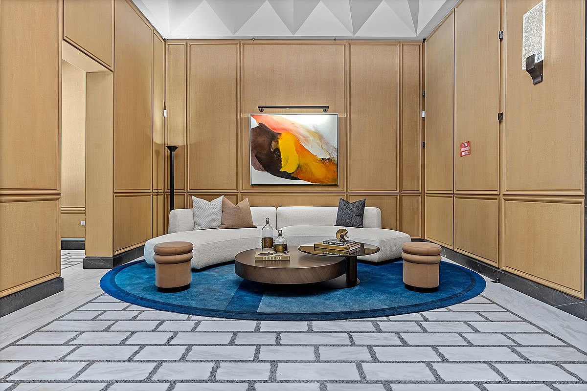 This sophisticated lobby or lounge area features a curved, cream-colored sofa centered on a vibrant blue circular rug. The space is defined by light wood-paneled walls with classic molding and a striking abstract painting illuminated by a picture light. A low, round coffee table sits in the middle, flanked by two small, cylindrical ottomans, all set against a patterned stone floor.