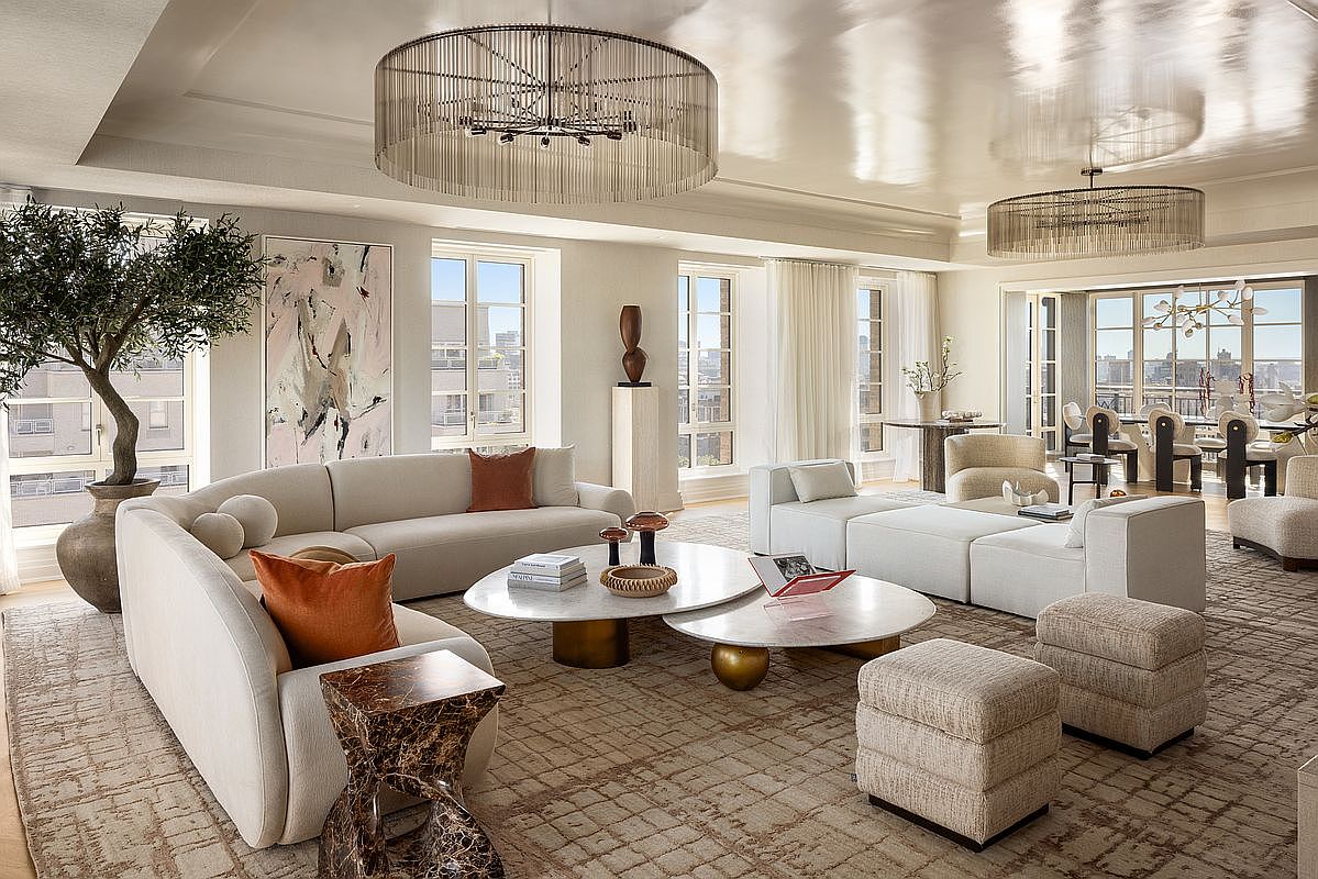 This expansive, light-filled living room features a sophisticated neutral palette with a large, curved sectional sofa and modular seating arrangements. The space is anchored by two overlapping circular stone-topped coffee tables and a textured area rug, while two dramatic, tiered fringe chandeliers hang from the high, reflective ceiling. Large windows provide ample natural light and views of the city, complemented by a potted olive tree and curated art pieces that enhance the elegant, contemporary aesthetic.
