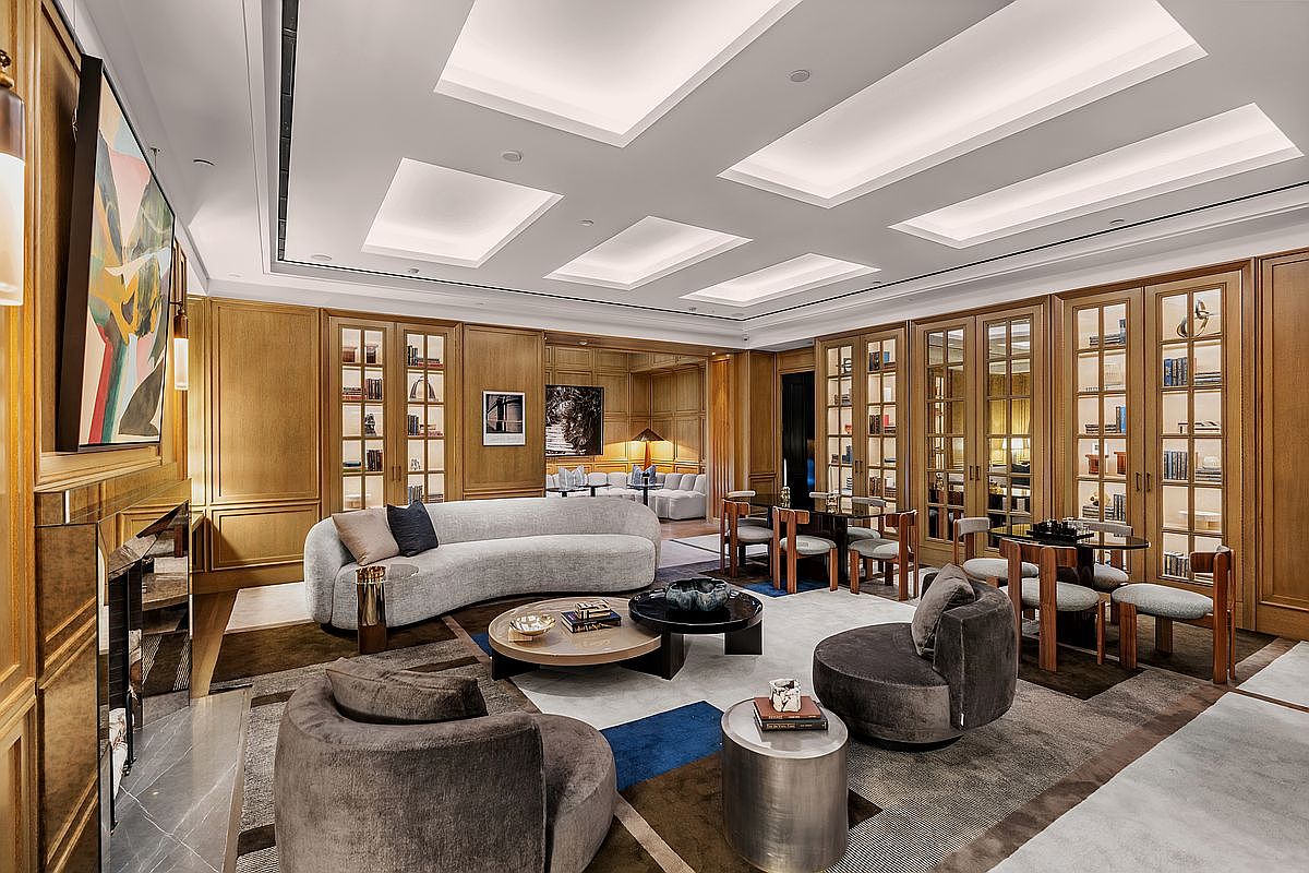 This sophisticated living room features warm wood-paneled walls with integrated glass-front cabinetry and a curved, light-toned sofa as the central focal point. The space is anchored by a layered area rug, a circular coffee table, and two plush, dark-toned armchairs, all set beneath a recessed ceiling with geometric lighting panels. The perspective captures a luxurious, open-plan lounge area that flows into a secondary seating space, exuding an atmosphere of refined elegance and comfort.